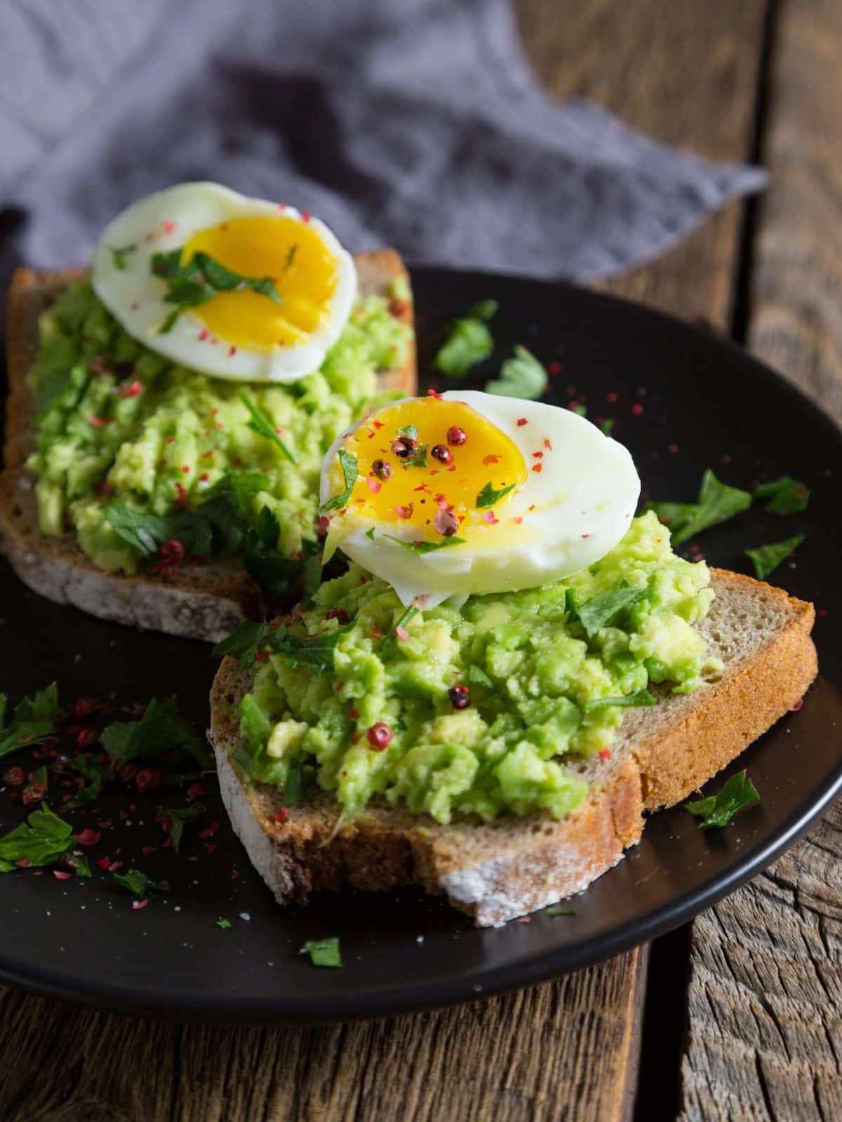 Avocado toast with a hard-boiled egg, red pepper flakes, and fresh herbs rests on a dark plate.