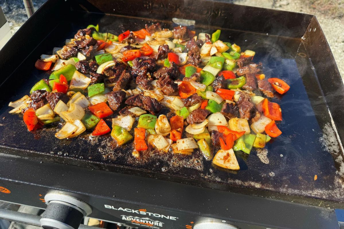 Sizzling mixed vegetables and meat chunks grilling to perfection on an outdoor flat top cooker.