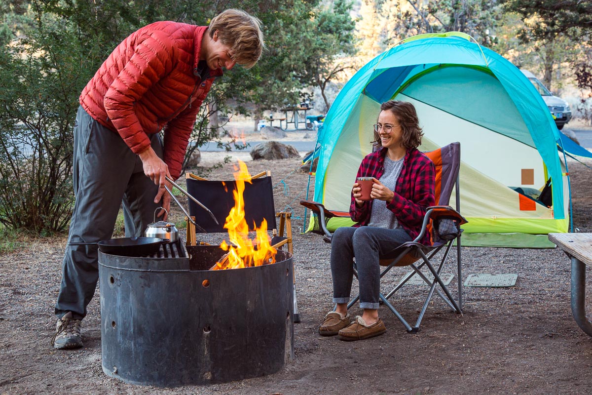 Two friends enjoying a camping trip with one tending to a campfire and the other sitting with a drink, sharing a moment of outdoor camaraderie.