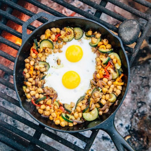 Chickpeas, vegetables, and two eggs in a skillet over a campfire.