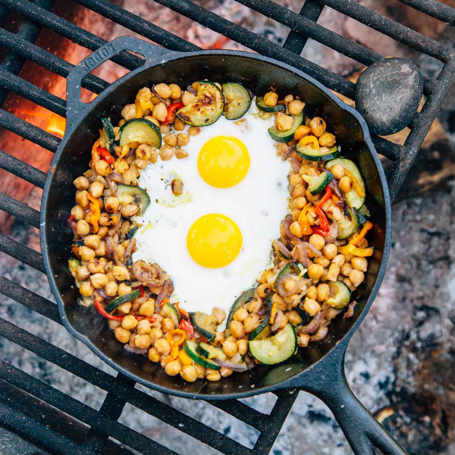 Chickpeas, vegetables, and two eggs in a skillet over a campfire.