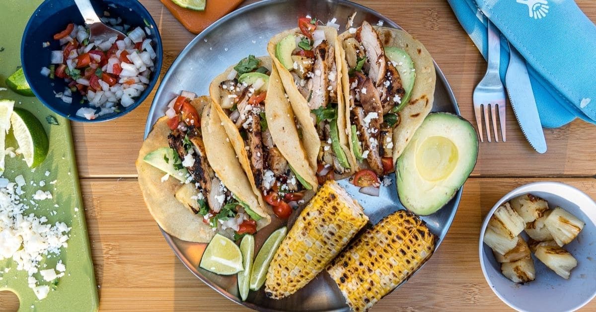 Four tacos arranged on a plate with grilled corn on the cob and hal an avocado