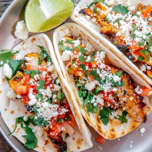 Three grilled chicken tacos on a plate with sliced limes