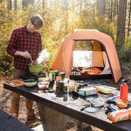 Michael standing at a camping table stirring tortellini soup in a pot. A camp scene is in the background