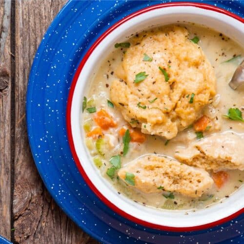 A bowl of creamy chicken and dumplings soup, garnished with herbs, sits on a blue speckled plate on a rustic wooden table with a spoon resting inside.