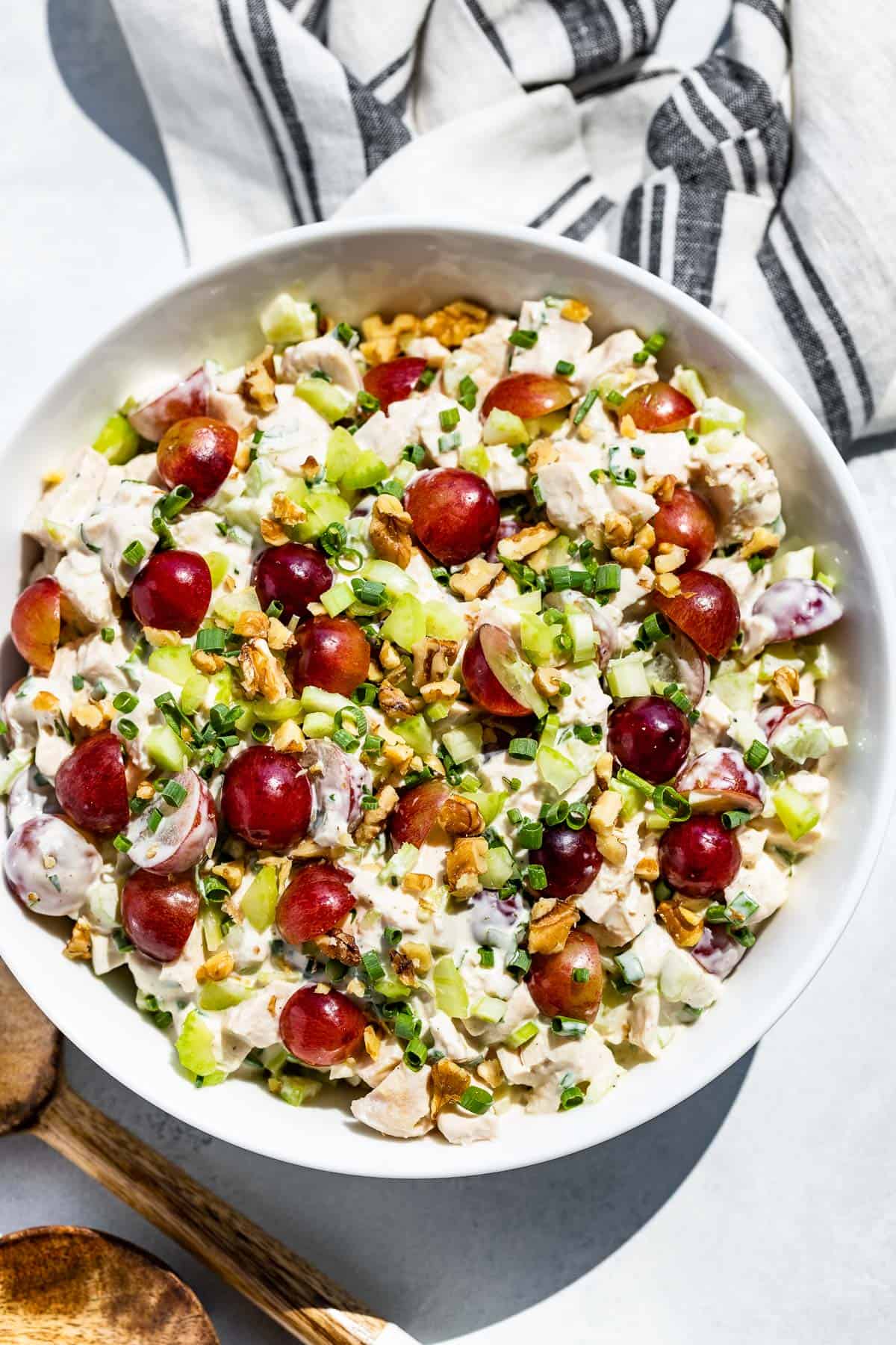 A colorful bowl contains chicken salad adorned with red grape halves, chopped celery, green onions, and walnuts.