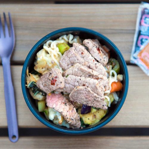 A bowl of noodle salad topped with grilled meat and vegetables, accompanied by a fork and condiment packets on a wooden table.