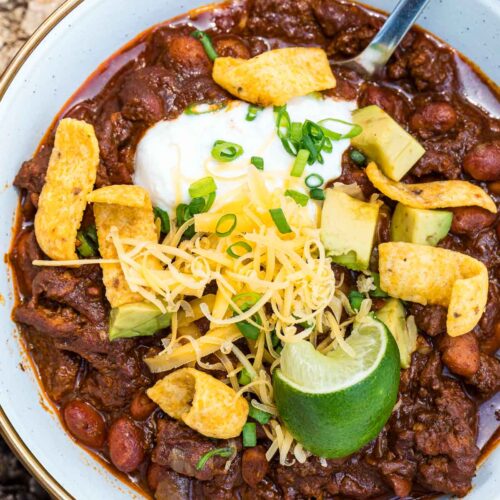 A bowl of chili topped with shredded cheese, diced avocado, sour cream, corn chips, and chopped green onions sits on a rustic wooden surface with a lime wedge on the side and a spoon tucked into the bowl.