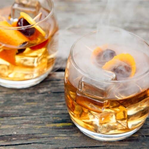 Two glasses of smoked old fashioned cocktails sit on a rustic wooden table with ice cubes, orange peels, cherries, and thin wisps of smoke rising from the drinks.