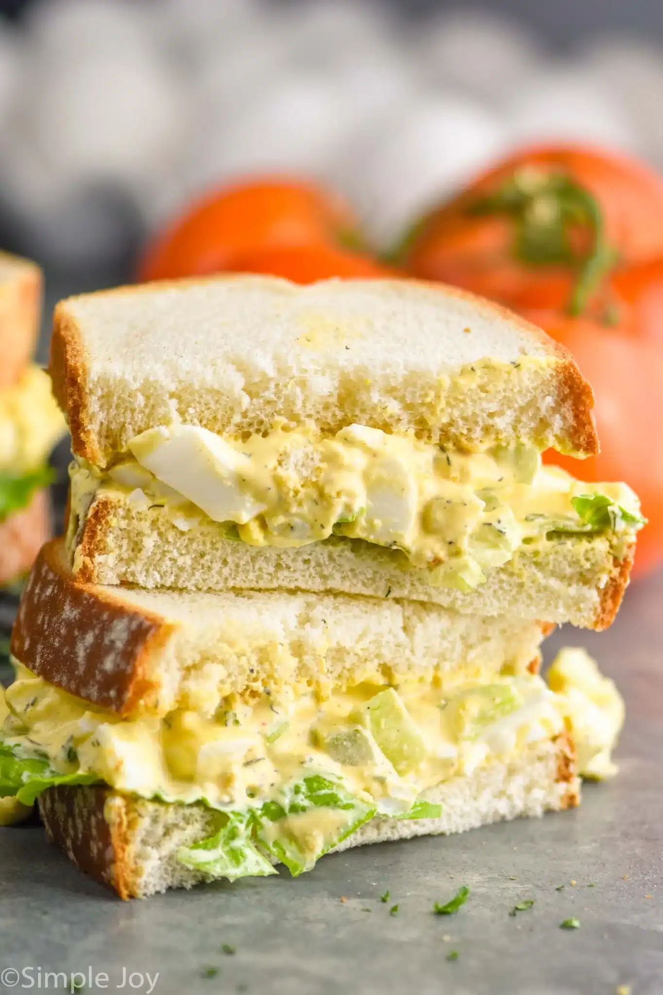 A freshly made egg salad sandwich with creamy filling, crisp lettuce, and soft bread is displayed close-up, surrounded by tomatoes.